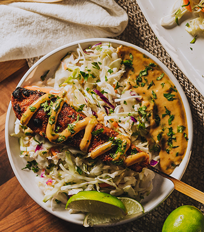 One turkey kabob rests on top of coleslaw and peanut sauce on a circular white plate. Wedges of lime garnish the dish.