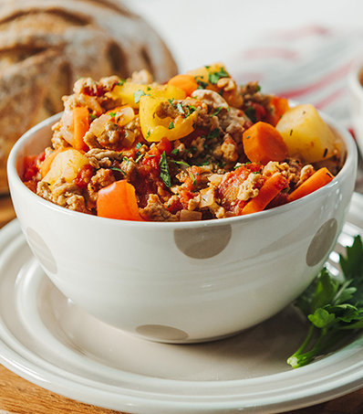 A serving of Bauerntopf, a German stew of ground pork, potatoes and carrots in a white bowl resting on a plate. A loaf of crusty bread can be seen out of focus behind the bowl of stew.
