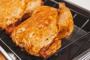 A whole freshly roasted chicken sits on a dark roasting rack and pan from the oven. A second chicken that is out of focus can be seen behind and slightly out of frame.