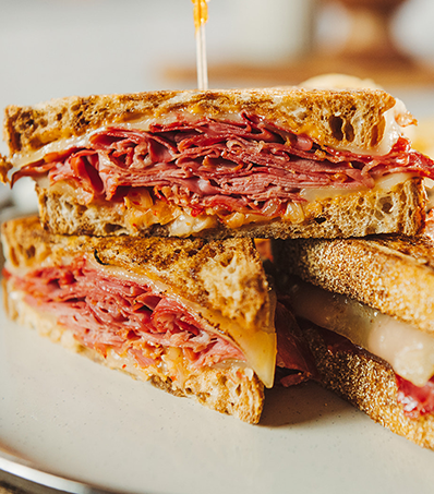 Three half sandwiches with layers of corn beef on rye bread sit on a light coloured plate. There are two half sandwiches on the bottom of the stack with one half on top with a took pick holding the stack together.