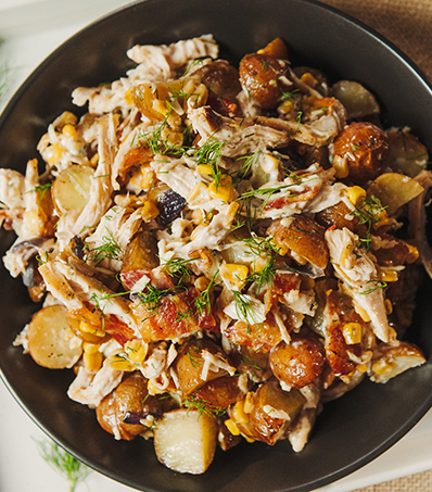 A top down view of a circular black bowl full of potato salad with torn pieces of cooked chicken and corn. The salad is garnished with fresh dill.