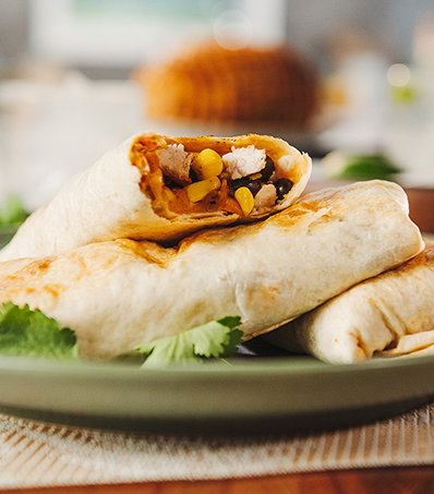 Three burritos rest on a green plate. The main focus is the top burrito stacked on the others with the corner of the tortilla missing to reveal the filling of cubed turkey, corn, black beans and peppers.